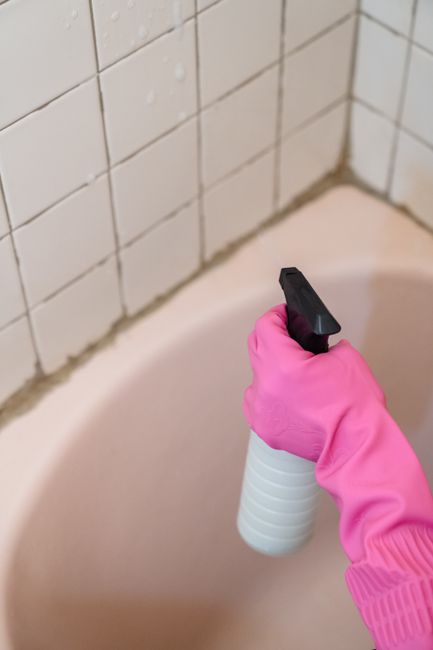 A person wearing a bright pink rubber glove is holding a white trigger spray bottle while applying cleaning solution to the grout of a bathroom bathtub. The bathtub is made of smooth, light pink mater